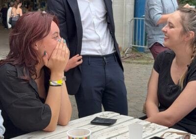 Goochelaar entertaint een groepje gasten aan tafel met een interactieve truc terwijl ze lachend en verrast reageren tijdens een buitenevent.