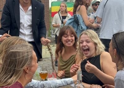 Goochelaar zorgt voor hilariteit met een interactieve truc terwijl een groepje vrienden luid lachend reageert aan een tafel tijdens een buitenevent.