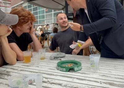 Goochelaar voert een interactieve kaarttruc uit aan een tafel terwijl een groepje vrienden aandachtig en nieuwsgierig toekijkt tijdens een buitenevent.