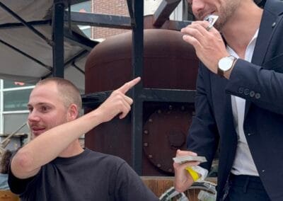 Goochelaar voert een verrassende kaarttruc uit en houdt een speelkaart in zijn mond terwijl een toeschouwer verbaasd reageert tijdens een buitenevent.