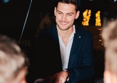 Goochelaar voert een interactieve kaarttruc uit aan tafel terwijl gasten toekijken tijdens een sfeervol avondfeest met verlichting op de achtergrond.