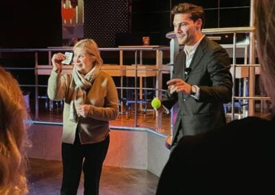 Goochelaar betrekt een toeschouwer bij een interactieve truc met een bal, terwijl een klein publiek toekijkt in een sfeervolle setting.